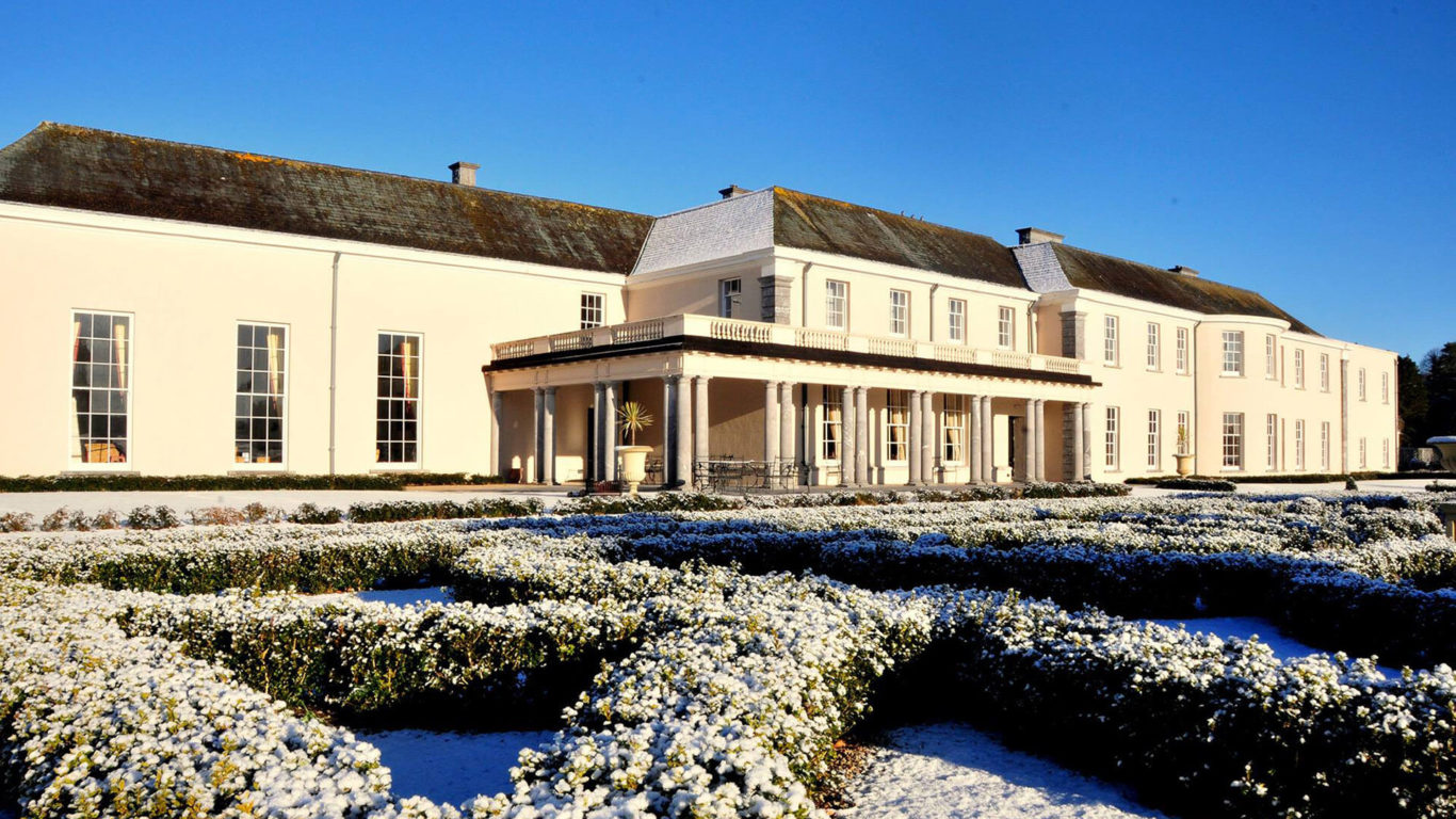 Elegant country hotel with snow-lined hedges, bright facade, and spacious grounds, inviting guests for a tranquil escape.