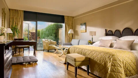 A well-lit hotel room with a double bed, sofa, desk, and glass doors leading to a private outdoor patio with greenery.
