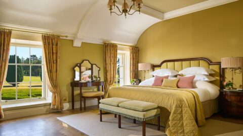 A spacious bedroom with a large bed, bench, vanity table, two windows with yellow curtains, yellow walls, and a chandelier, overlooking a garden.