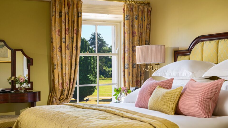A neatly made bed with pastel pillows and a yellow blanket sits beside a window with patterned curtains, overlooking a green garden. A lamp and flowers are on the bedside table.