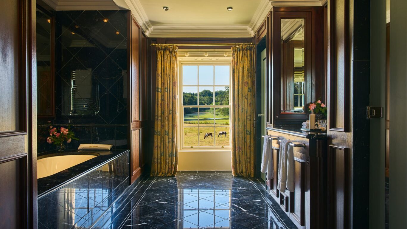Luxurious bathroom with black marble floor and bathtub, wood paneling, large window with yellow curtains, and a view of a green field with horses outside.
