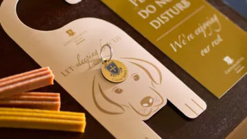 A close-up of a hotel room door hanger with a dog illustration, a keychain, and a “Please Do Not Disturb” sign on a table, with dog treats nearby.