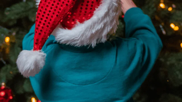 A child wearing a red Santa hat decorates a Christmas tree with lights and ornaments.