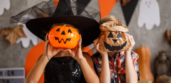 Two children in Halloween costumes hold pumpkin-themed containers over their faces, with festive decorations like paper ghosts and pumpkins in the background.