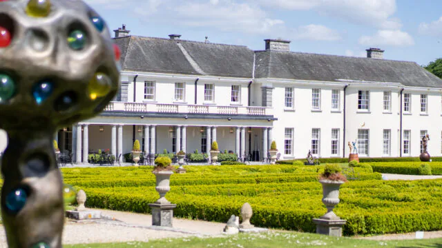 Elegant white hotel with classic columns, lush gardens, art sculptures, under a bright blue sky, offering tranquil relaxation.