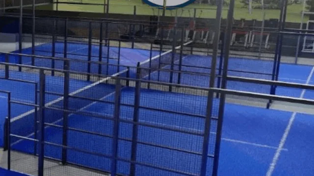 Indoor padel tennis courts with blue flooring and black metal fences under a wooden roof, with a