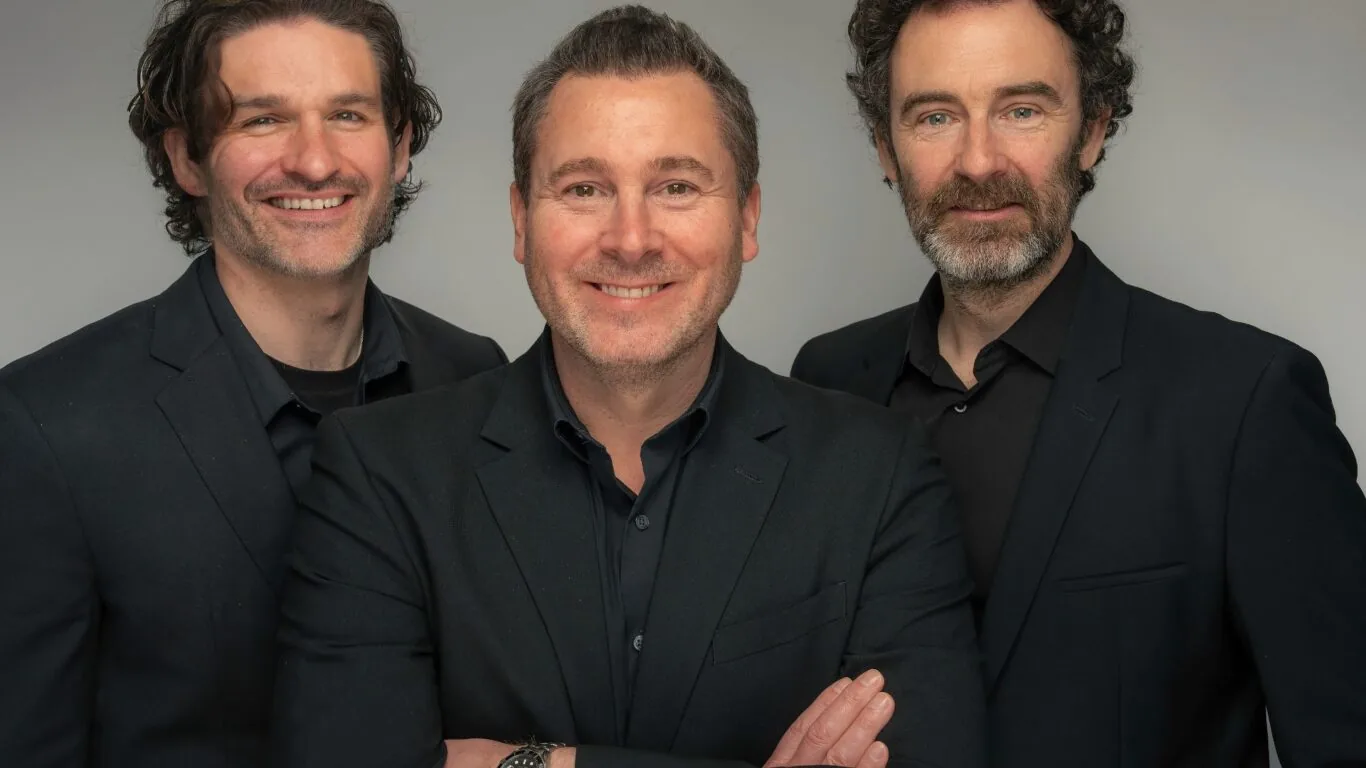 Three men in black suits and dark shirts stand together against a plain background, smiling and looking at the camera.