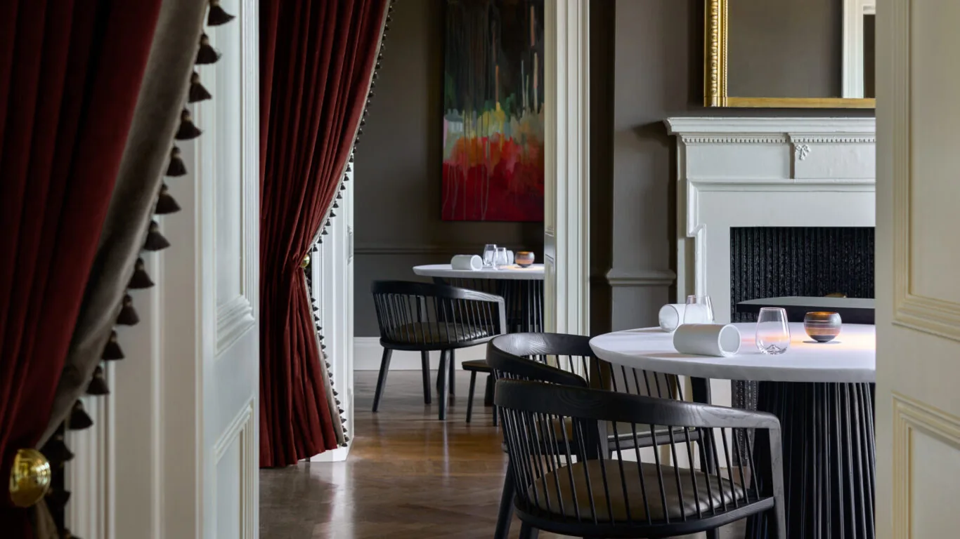 Elegant dining room with round tables, modern black chairs, a fireplace, dark red curtains, and abstract artwork on the wall. Table settings are minimal and the space appears quiet.