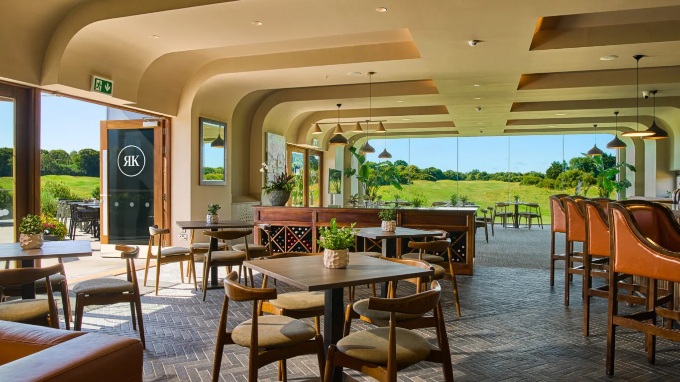Modern restaurant interior with wooden tables and chairs, leather seating, and large windows overlooking a green outdoor landscape.