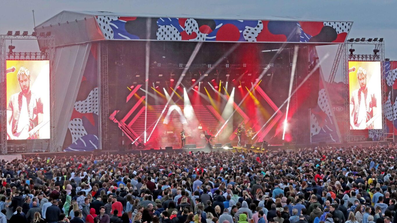 Large crowd gathered in front of a colorful outdoor concert stage with bright lights and video screens showing a performer playing guitar.