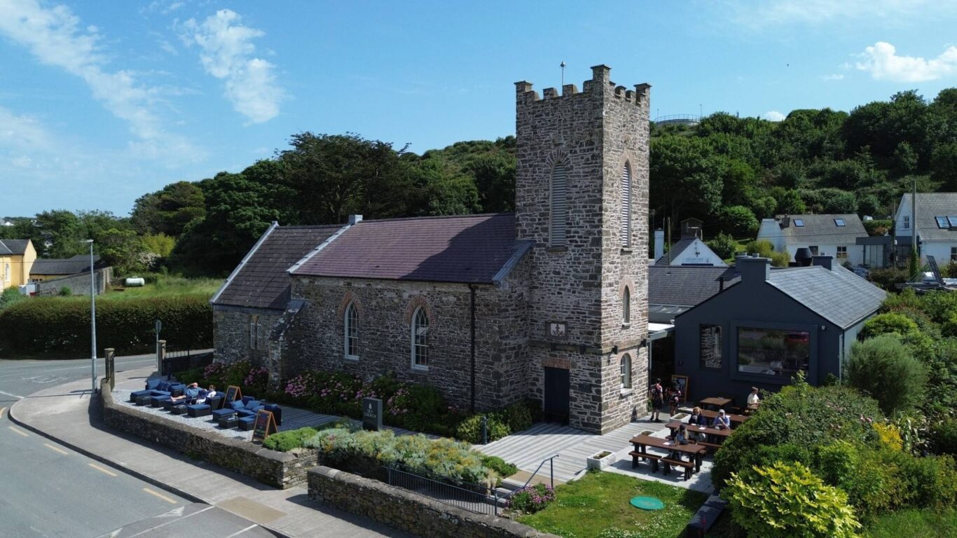 A stone church building with a square tower is surrounded by seating areas, greenery, and nearby houses on a sunny day.