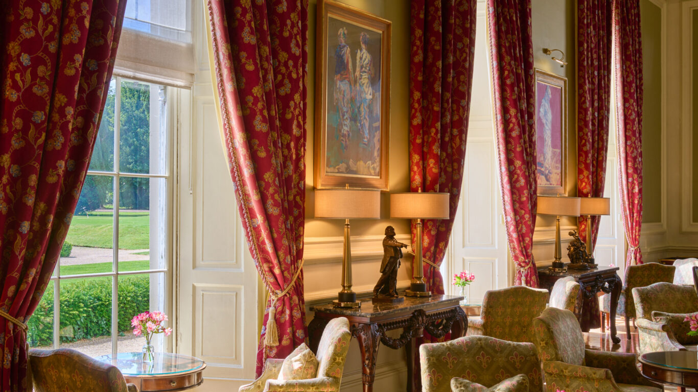 A spacious, elegant sitting room with tall windows, red patterned curtains, armchairs, small tables with flowers, and decorative lamps on wooden side tables under framed art.