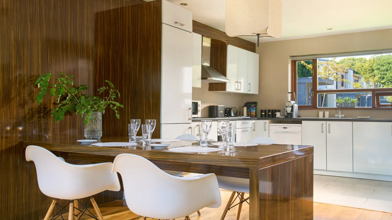 Modern kitchen and dining area with wood paneling, white cabinets, four white chairs, a rectangular table set with glassware, and large window letting in natural light.