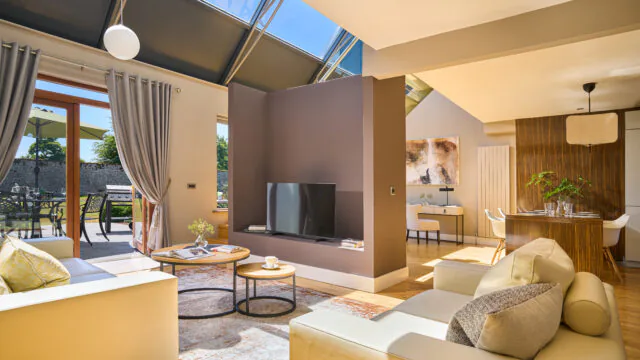 Spacious, sunlit hotel suite with a cosy lounge, modern decor, large windows, and patio access, ideal for relaxation.