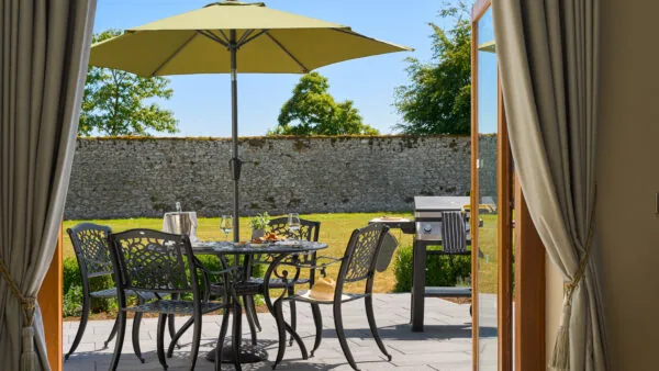 A patio with a metal table and chairs under a green umbrella, a barbecue grill nearby, and a stone wall in the background on a sunny day.