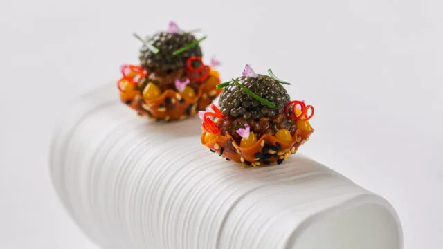 Two intricately plated hors d’oeuvres garnished with herbs and caviar are displayed on a curved, white porcelain stand set on a white tablecloth.