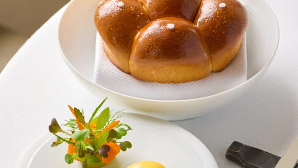 A plate of golden brown dinner rolls sits beside a smaller plate with a quenelle of butter and a garnish of microgreens on a white tablecloth.