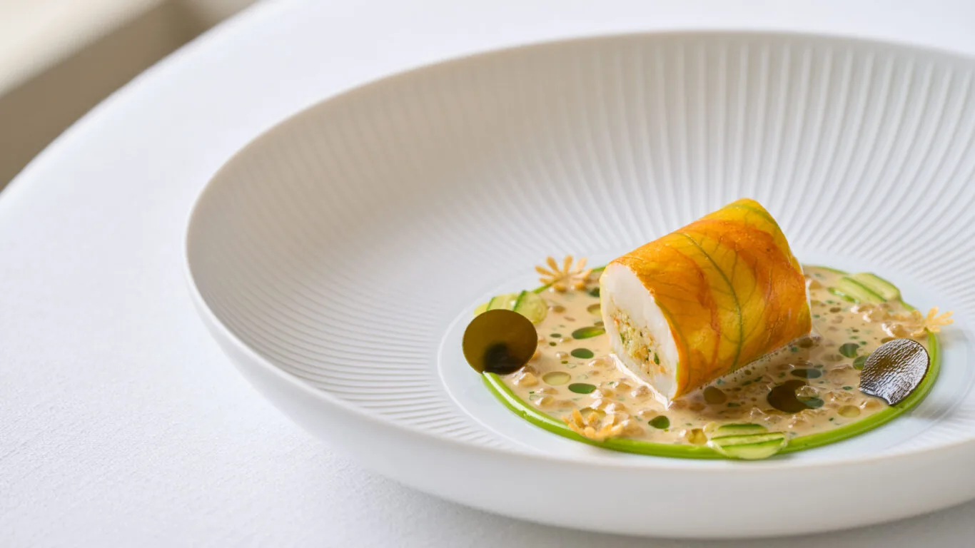 A plated gourmet dish featuring a rolled protein atop a creamy grain mixture, garnished with green sauce, thin vegetable slices, and small edible flowers, served in a white bowl.
