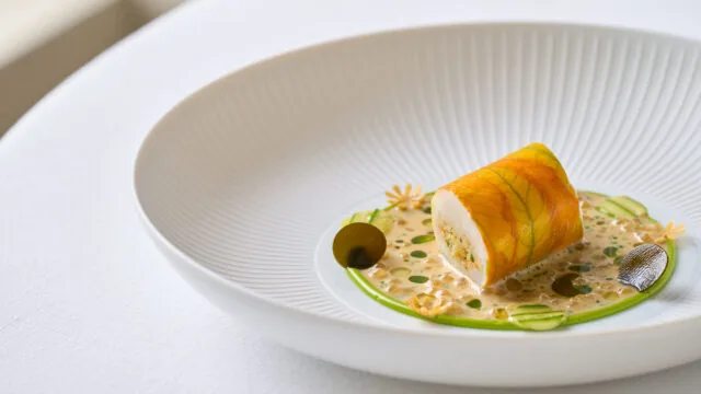 A plated gourmet dish featuring a rolled protein atop a creamy grain mixture, garnished with green sauce, thin vegetable slices, and small edible flowers, served in a white bowl.