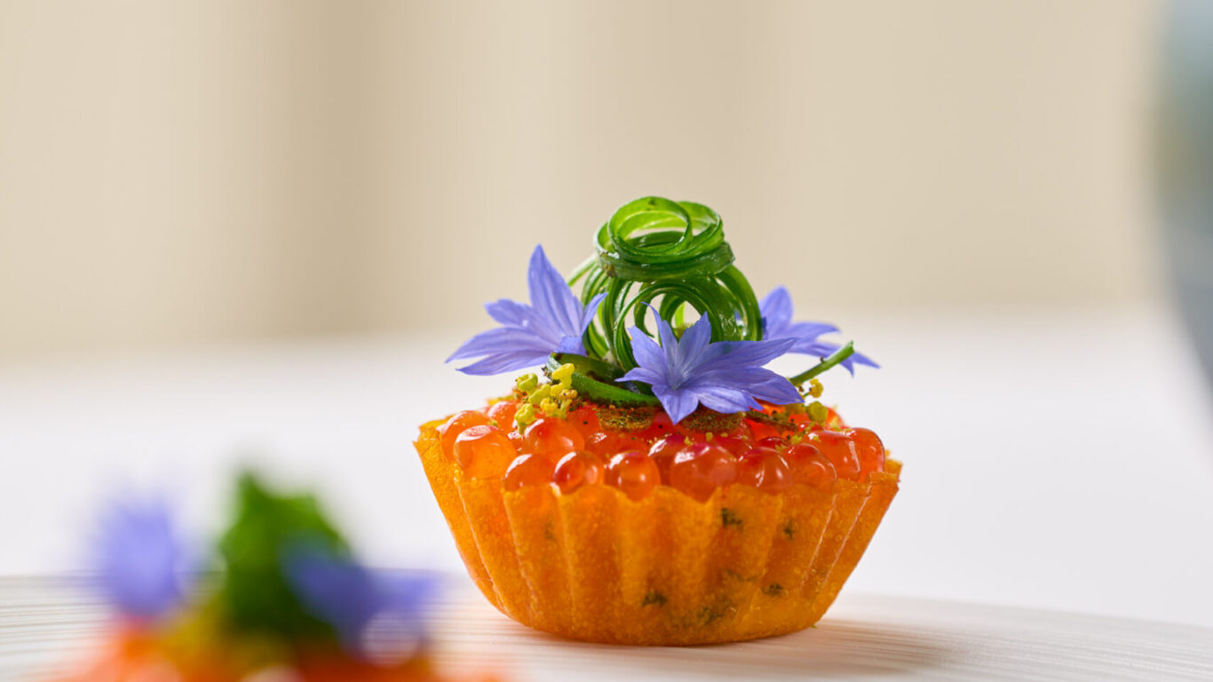 A small tart topped with orange caviar, green garnish, and purple edible flowers on a white plate.