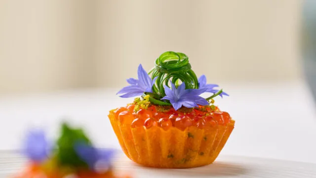 A small tart topped with orange caviar, green garnish, and purple edible flowers on a white plate.