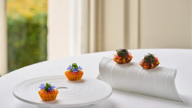 Three bite-sized appetizers with colorful garnishes are presented on modern white ceramic serving dishes, placed on a white tablecloth near a window.