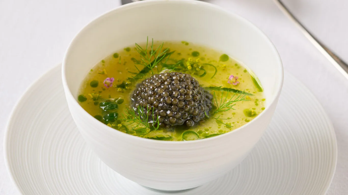 A white bowl of green soup garnished with a mound of caviar and herbs, placed on a matching plate with a spoon and knife in the background.