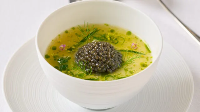 A white bowl of green soup garnished with a mound of caviar and herbs, placed on a matching plate with a spoon and knife in the background.