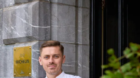 A chef in a white uniform stands with arms crossed in front of a building with a Michelin plaque on the wall.