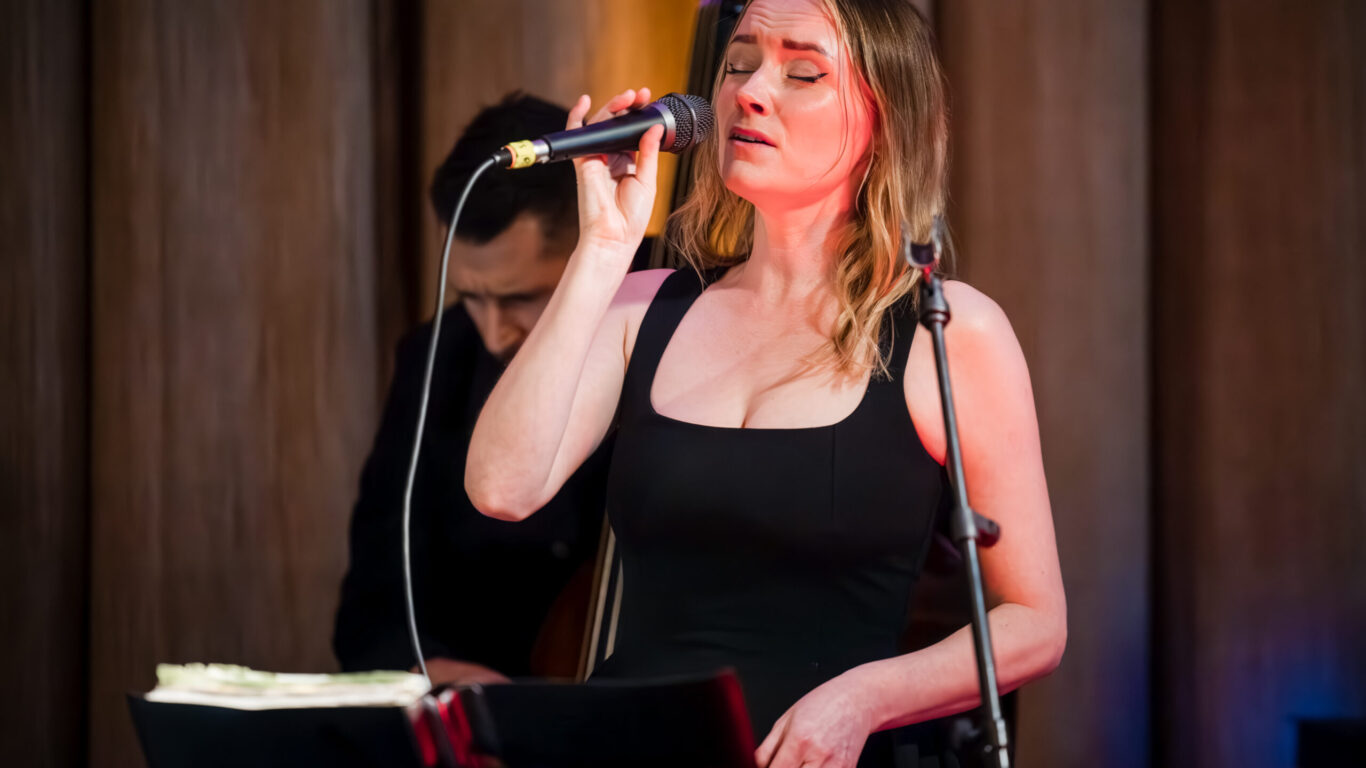 A woman in a black dress sings into a microphone on stage, with a man playing an upright bass in the background.