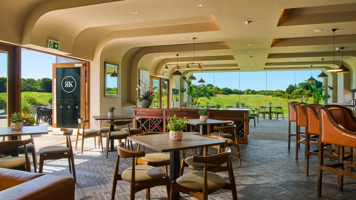 Modern restaurant interior with wooden tables and chairs, large windows offering views of green fields, and natural light filling the space.