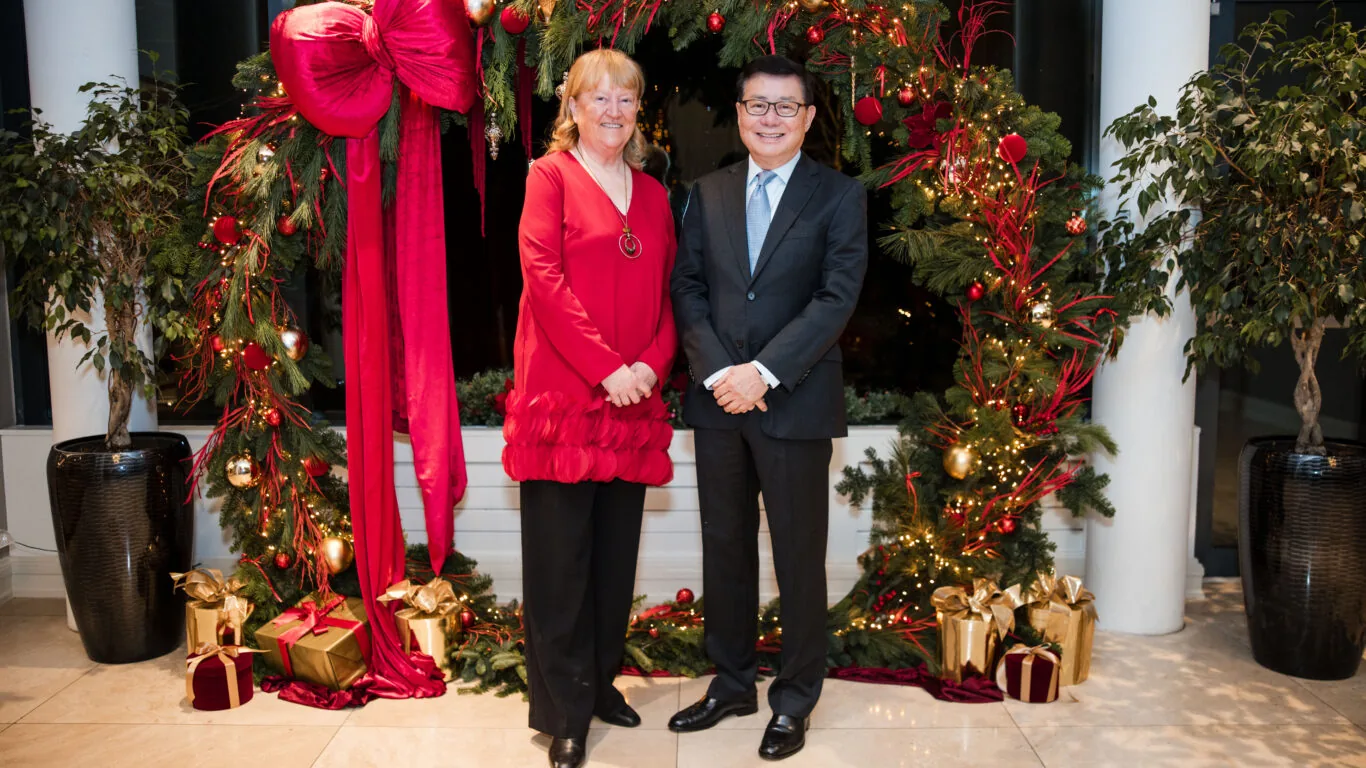 Two people stand in front of a large, decorated Christmas wreath with red ornaments, bows, and wrapped gifts on the floor. Both are dressed formally and smiling at the camera.