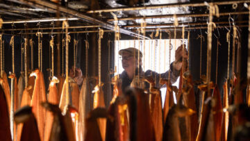 A person stands among rows of fish fillets hanging from hooks inside a smokehouse, with light streaming in from a doorway behind them.