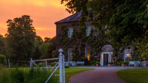A large, ivy-covered country house with arched windows and outdoor seating, set against a sunset sky and surrounded by trees and greenery.