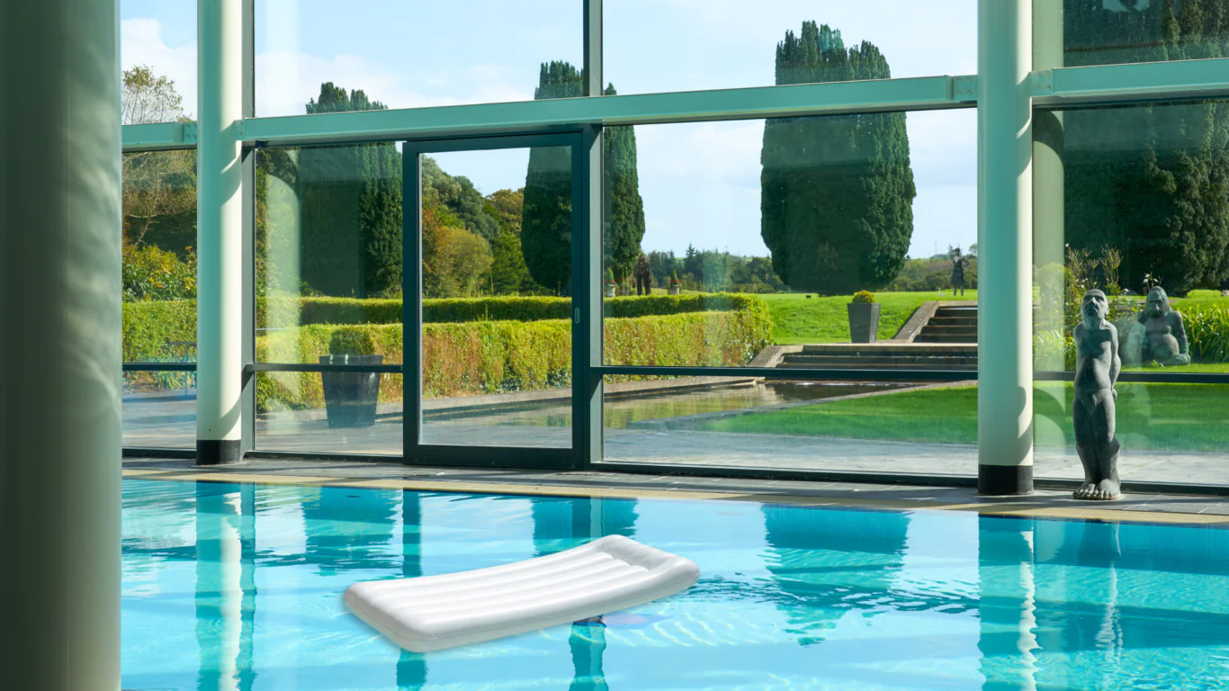 Luxurious indoor pool with garden view, featuring a white lounger and elegant sculptures, offering serene relaxation.