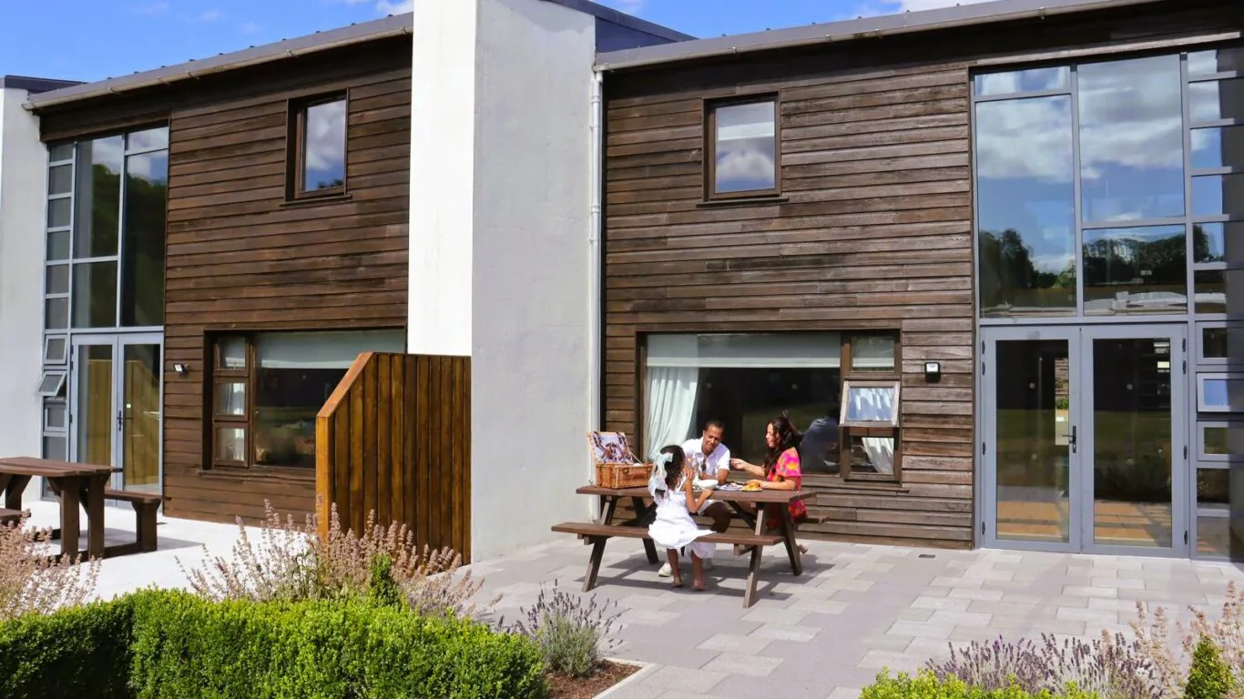 Guests enjoying a sunny day at a modern wooden hotel, sitting at an outdoor table surrounded by greenery.