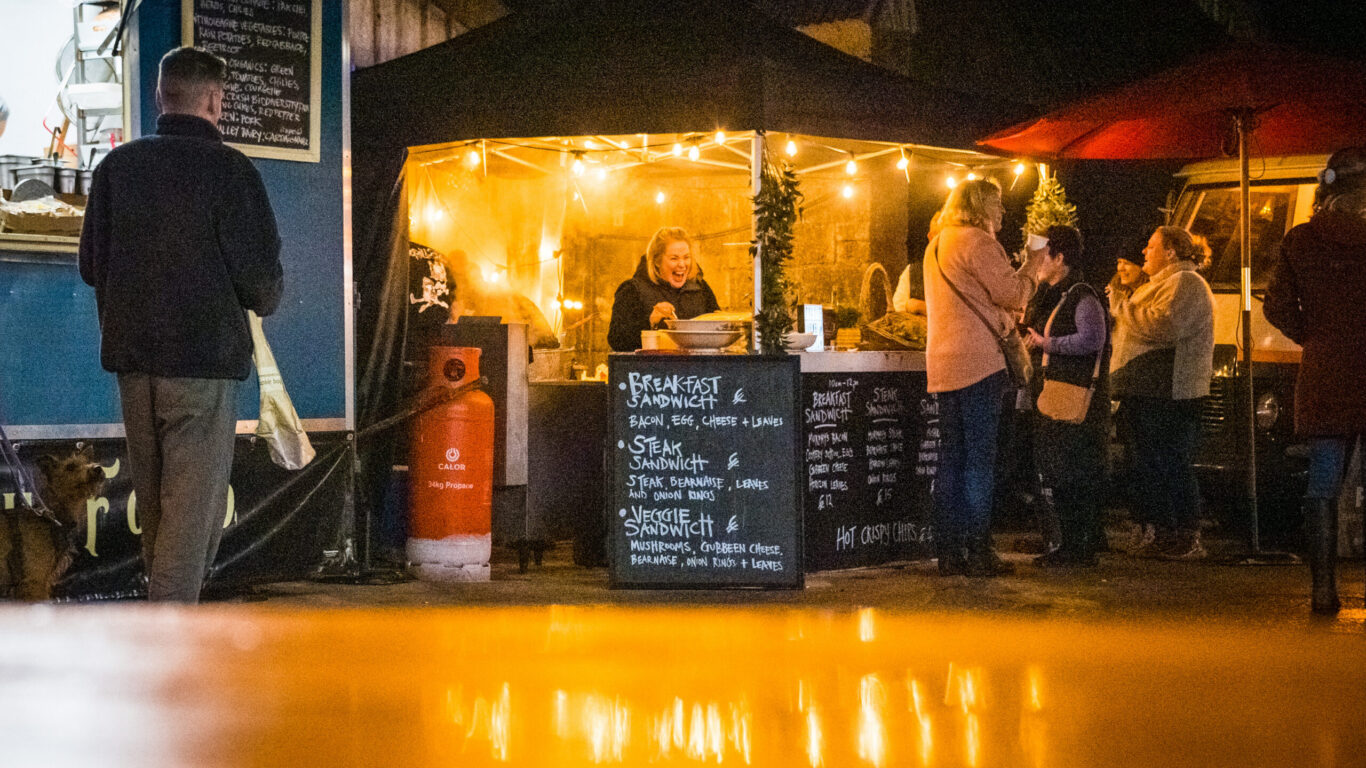 Guests enjoy a cosy evening at a food stall with warm lights, offering breakfast sandwiches at the hotel&rsquo;s outdoor market.