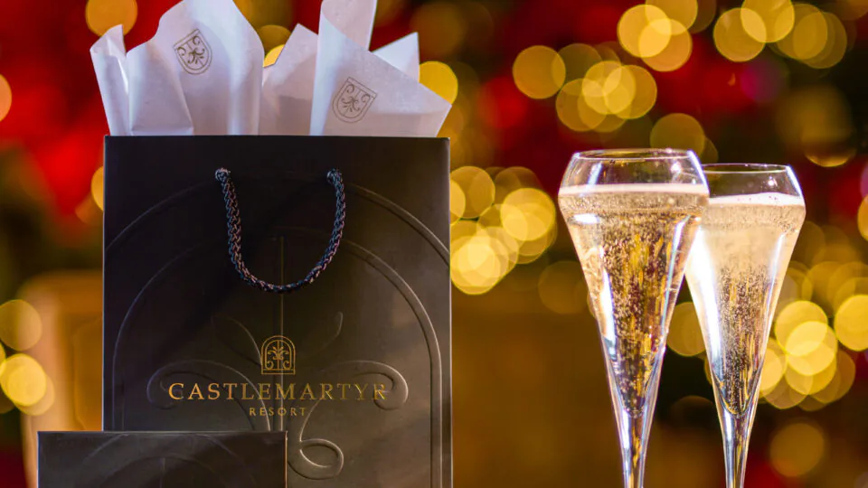 Luxurious gift bag and sparkling wine glasses at Castlemartyr Resort with festive gold bokeh lights.